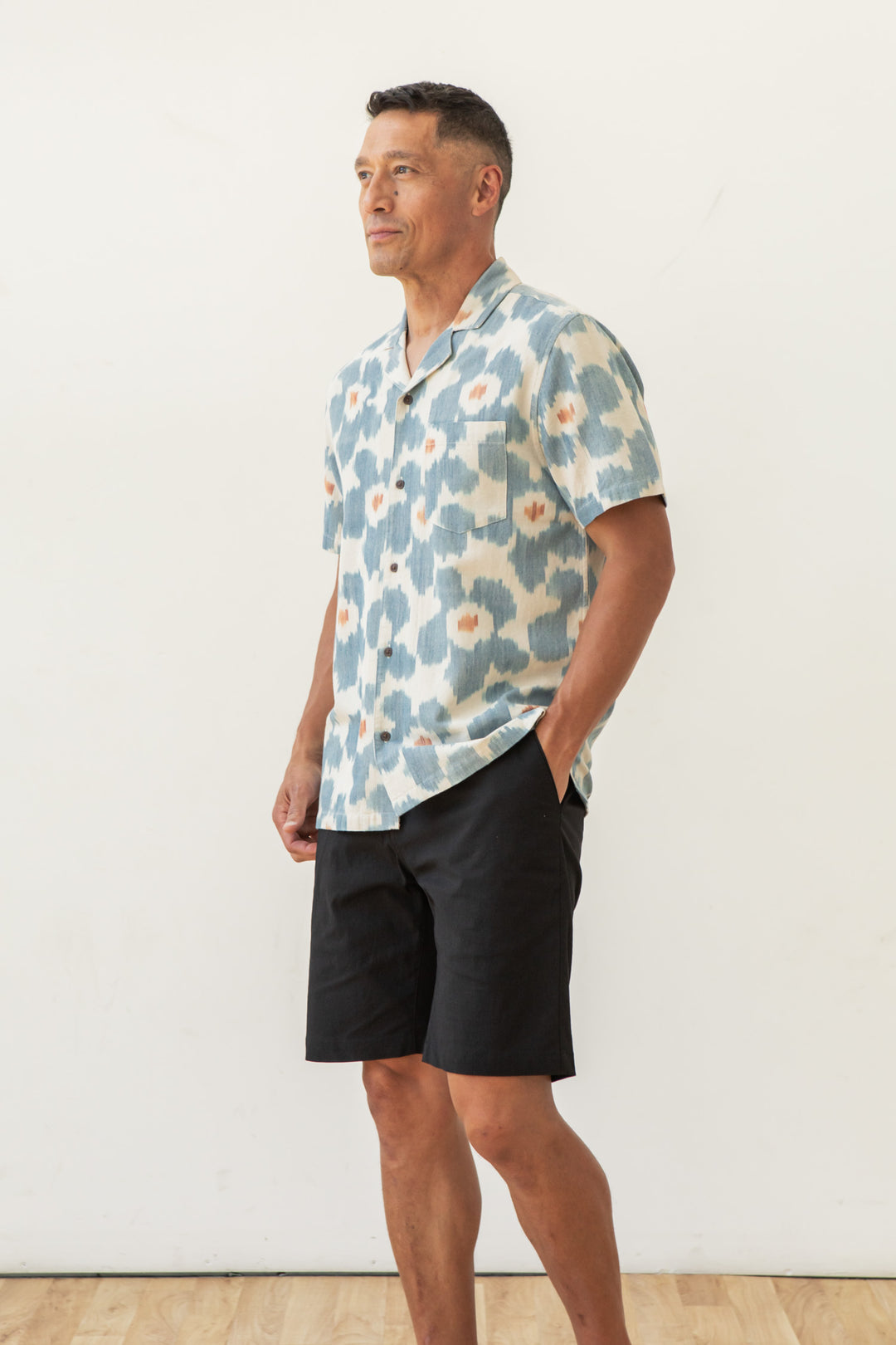 A man stands against a plain white background, wearing the Bridge & Burn Bodie Shirt in Polar Blossom, featuring blue and white patterns, paired with black shorts. One hand is in his pocket as he looks to the side with a smile.