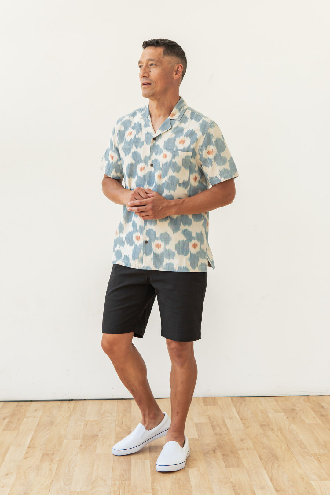 A man stands indoors on a wooden floor, wearing the Bridge & Burn Bodie Shirt in Polar Blossom, black shorts, and white slip-on shoes, posed against a plain white wall.