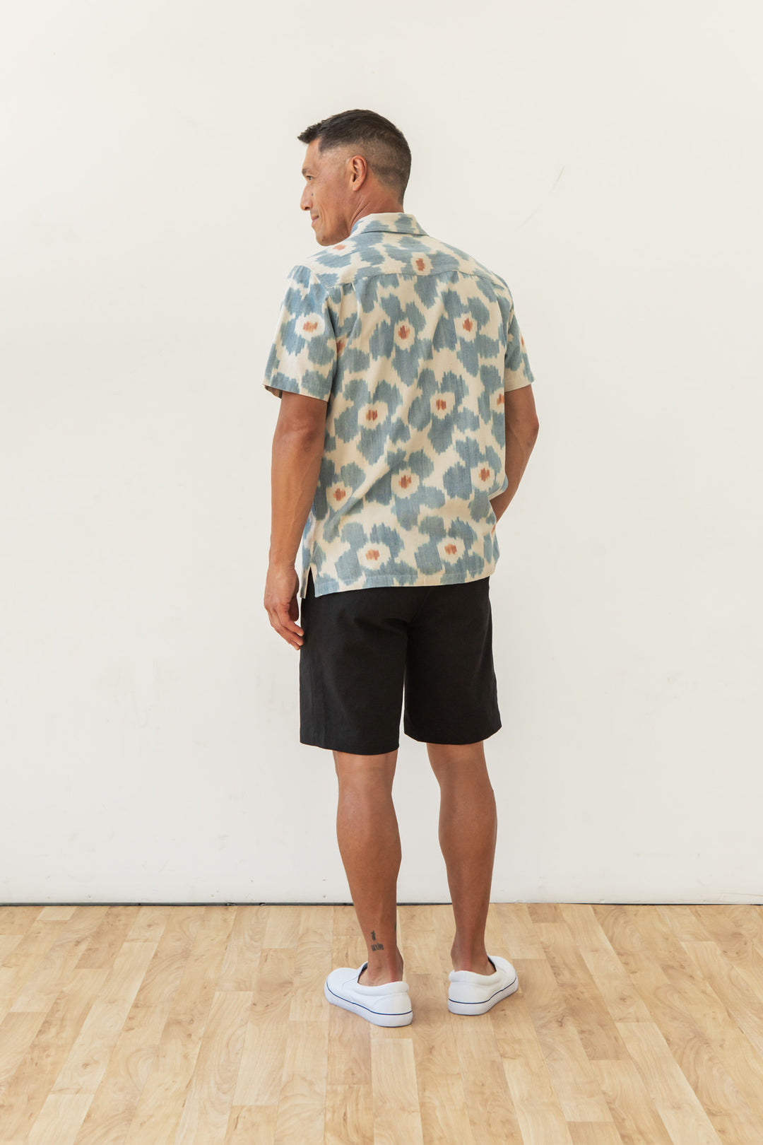 A man stands on a wooden floor, facing away, wearing Bridge & Burn's Bodie Shirt in Polar Blossom with blue and white florals, paired with black shorts and white slip-on shoes against a plain white wall.