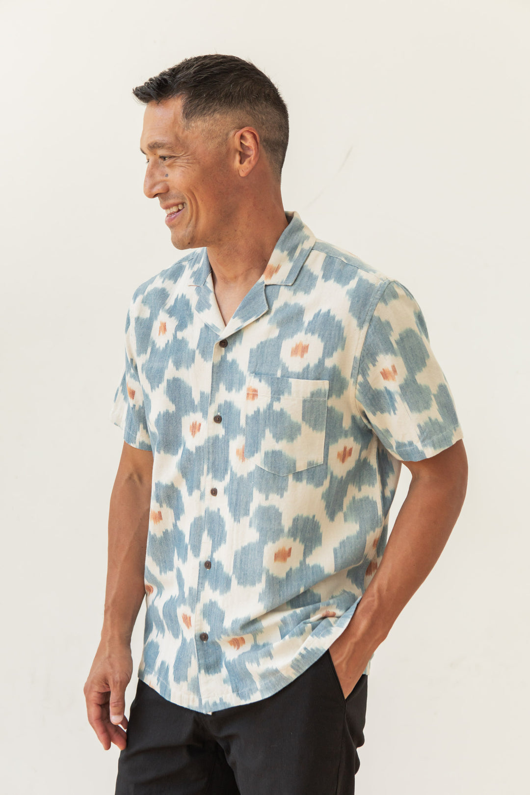 A man wears the Bridge & Burn Bodie Shirt in Polar Blossom, a relaxed fit with a blue and white abstract floral pattern, smiling and looking to his left against a plain light-colored background.