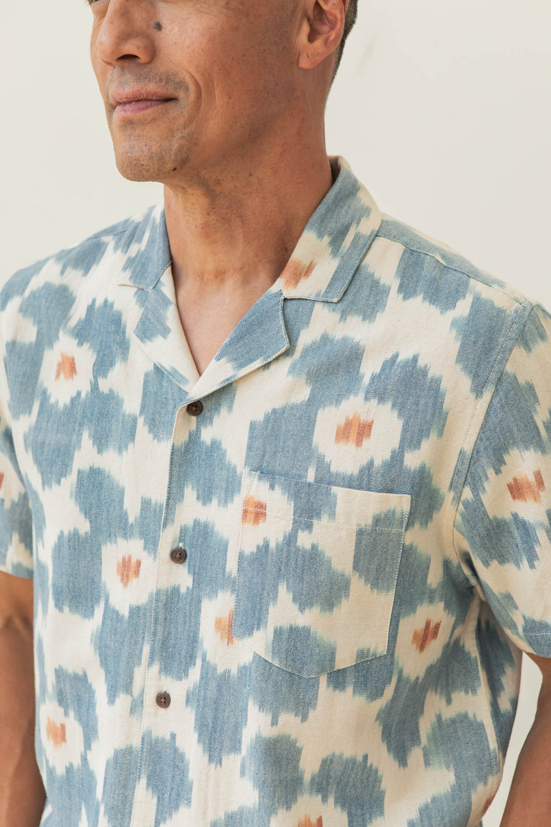 A man wears the Bridge & Burn Bodie Shirt in Polar Blossom, a relaxed fit summer cotton shirt with a blue and white abstract pattern, orange accents, retro camp collar, chest pocket, and brown buttons.