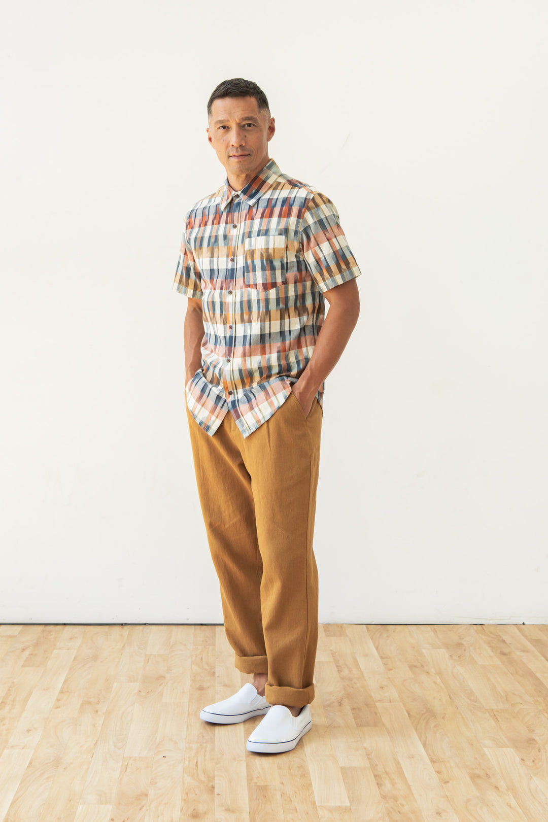 A man stands on a wooden floor against a plain white wall, wearing the Bridge & Burn Harbor Slim Shirt in Aurora Space Dye Plaid, tan trousers, and white slip-on shoes, with his hands in his pockets for an effortlessly stylish look.