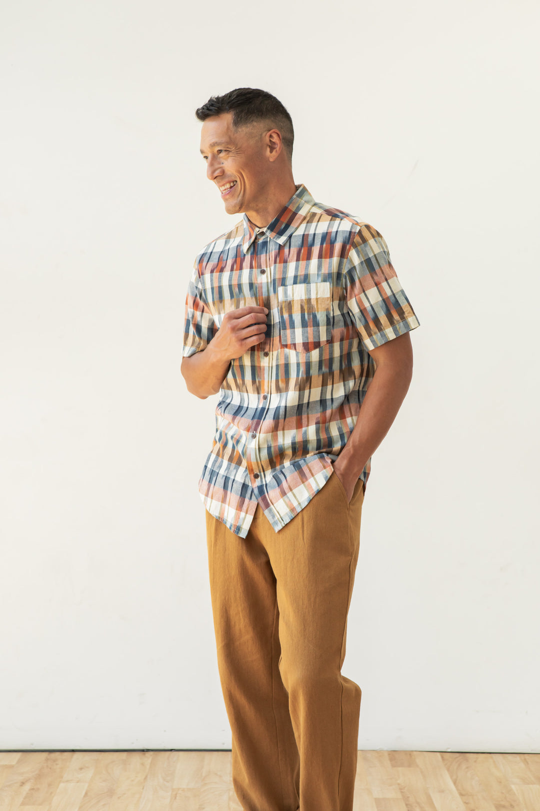 A man smiles against a white background, one hand in his pocket and the other on his chest, wearing the Bridge & Burn Harbor Slim Shirt in Aurora Space Dye Plaid with tan pants—showcasing this shirt’s versatile, slim fit style.