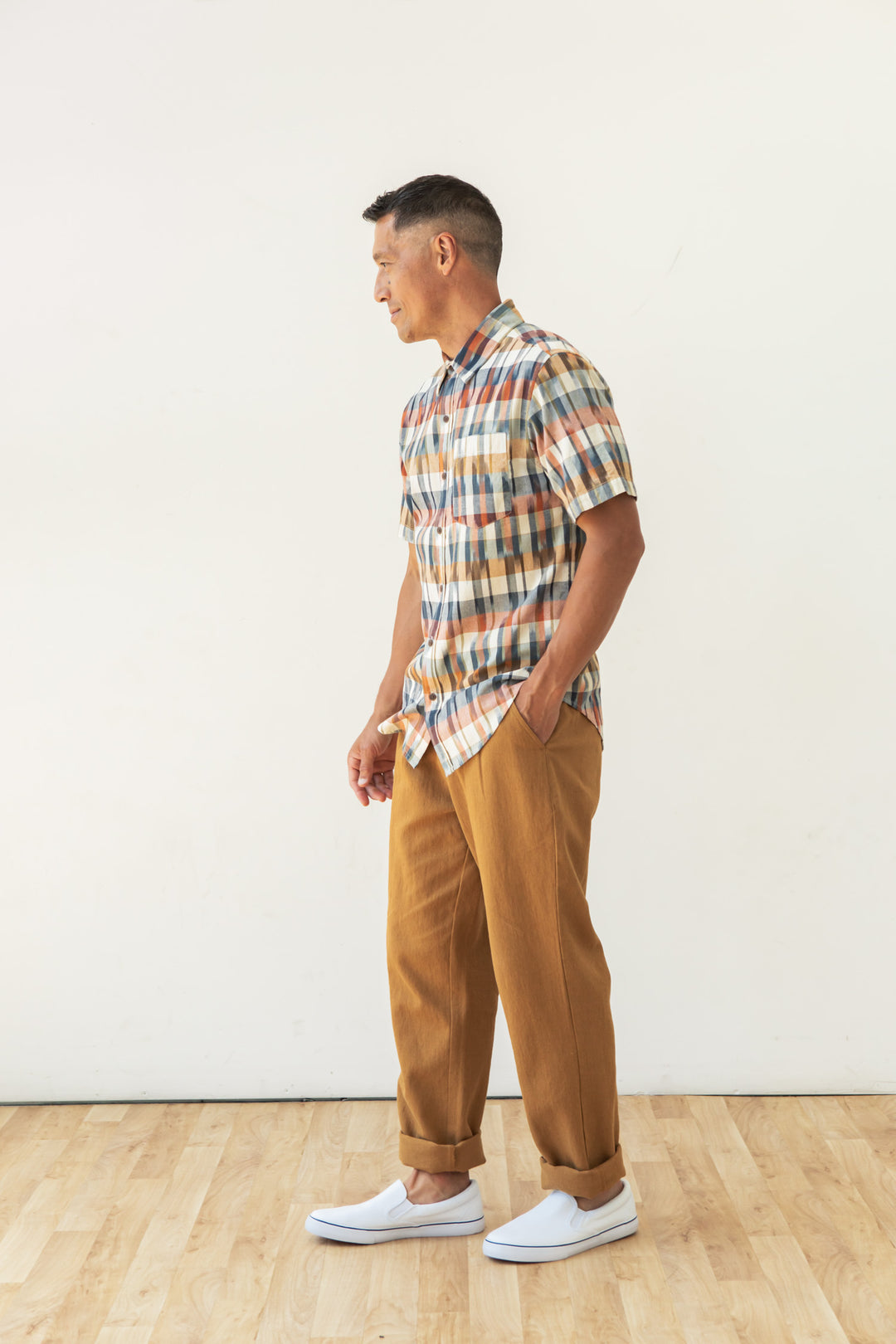 A man stands on a wooden floor against a white wall, wearing the Bridge & Burn Harbor Slim Shirt in Aurora Space Dye Plaid, tan pants, and white slip-on shoes, with one hand in his pocket and looking to the side.