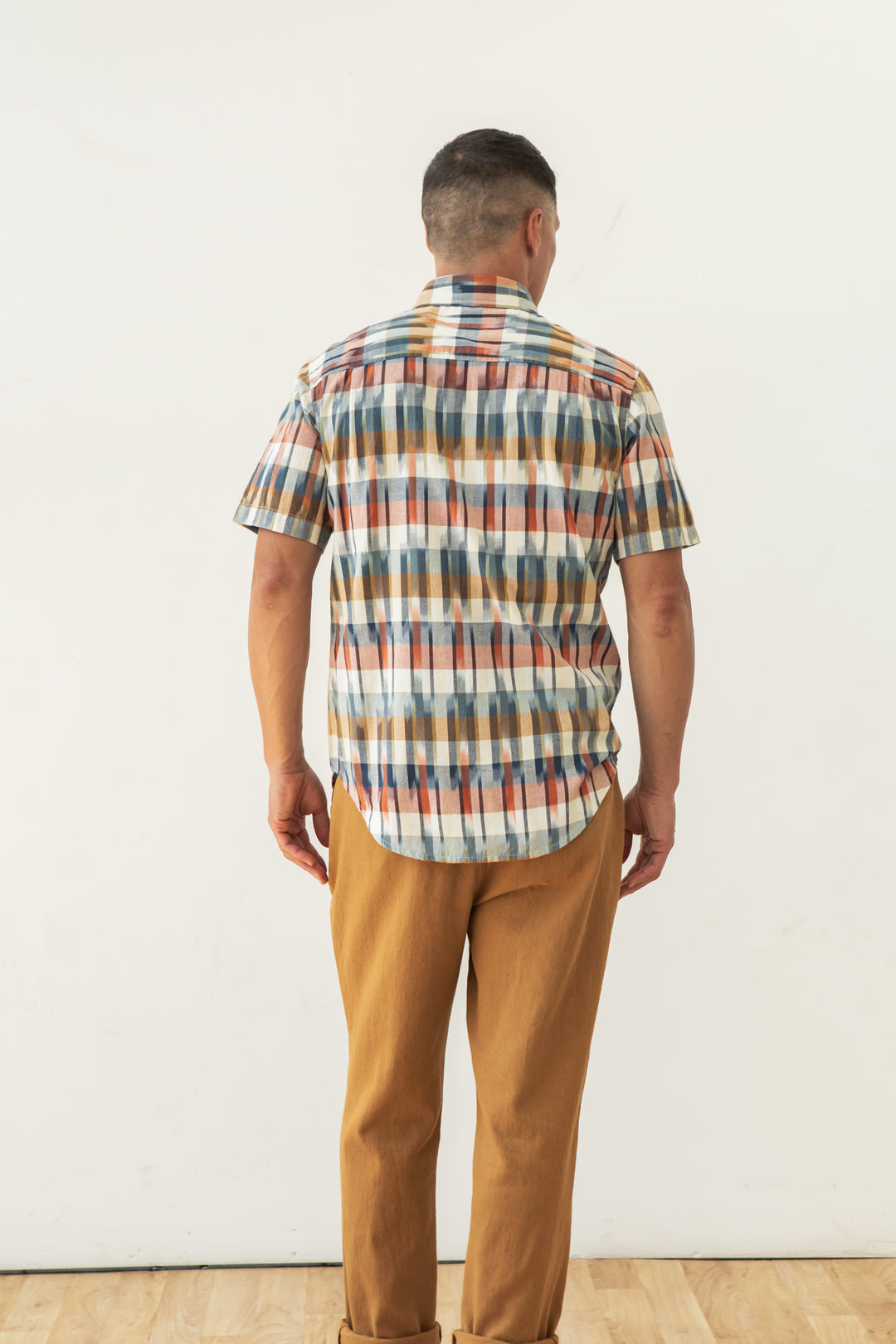 A man faces away from the camera, wearing the Bridge & Burn Harbor Slim Shirt in Aurora Space Dye Plaid and light brown pants, set against a plain white background.