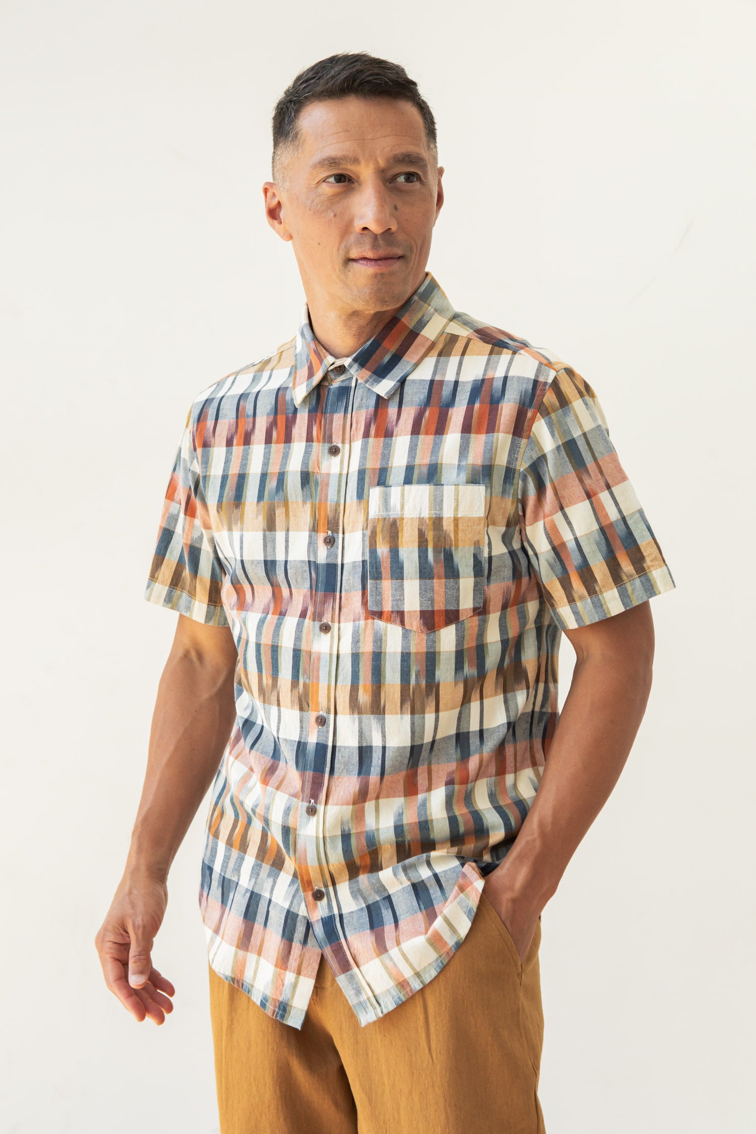 A man stands against a plain, light background in the Bridge & Burn Harbor Slim Shirt in Aurora Space Dye Plaid, short-sleeved and multicolored, paired with light brown pants. He looks slightly to the side with one hand in his pocket.
