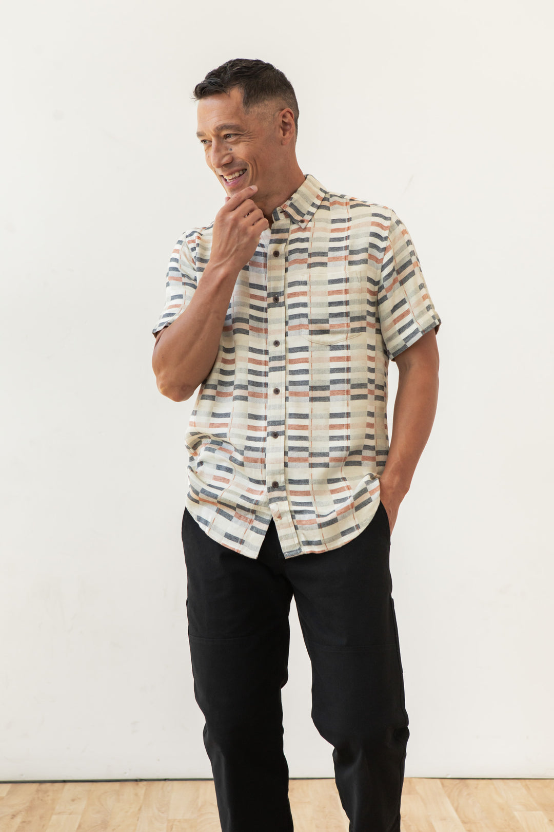 A man smiles against a white background, wearing the Bridge & Burn Jordan Slim Shirt in Broadway Stripe with black pants. One hand is in his pocket, the other touches his chin—showcasing this modern wardrobe essential.