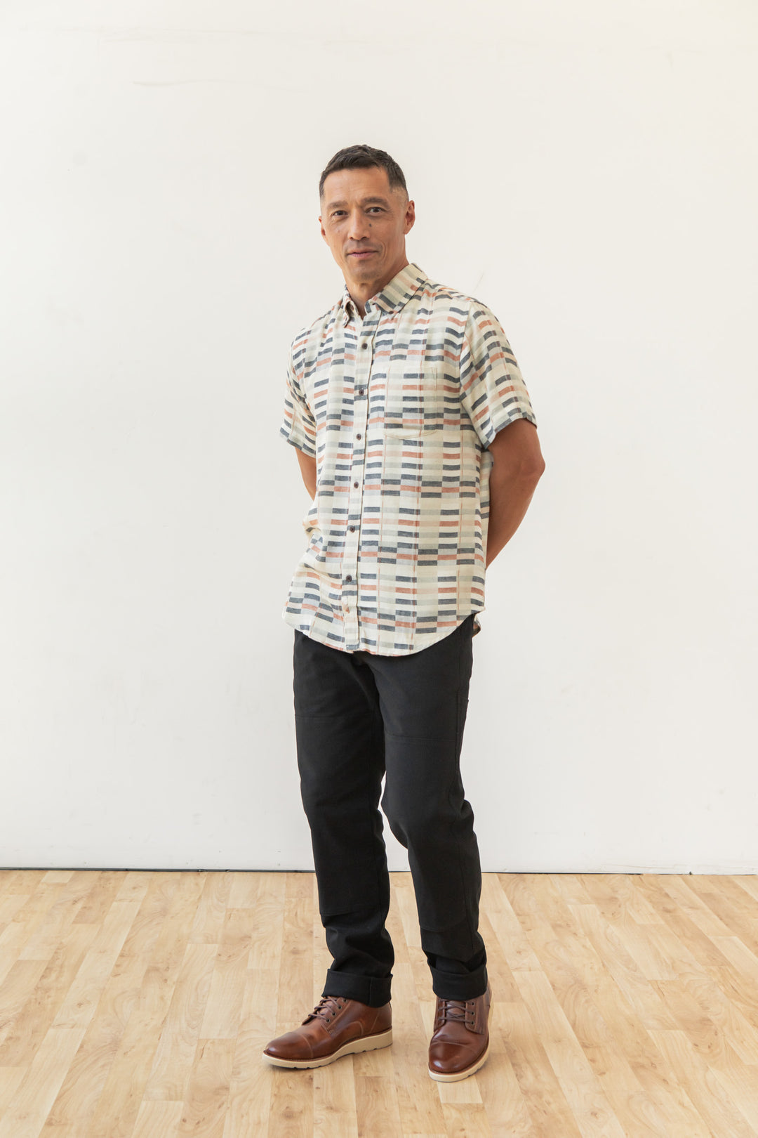 A man stands indoors on a wood floor against a plain white wall, wearing the Bridge & Burn Jordan Slim Shirt in Broadway Stripe, paired with black pants and brown boots, his hands behind his back.