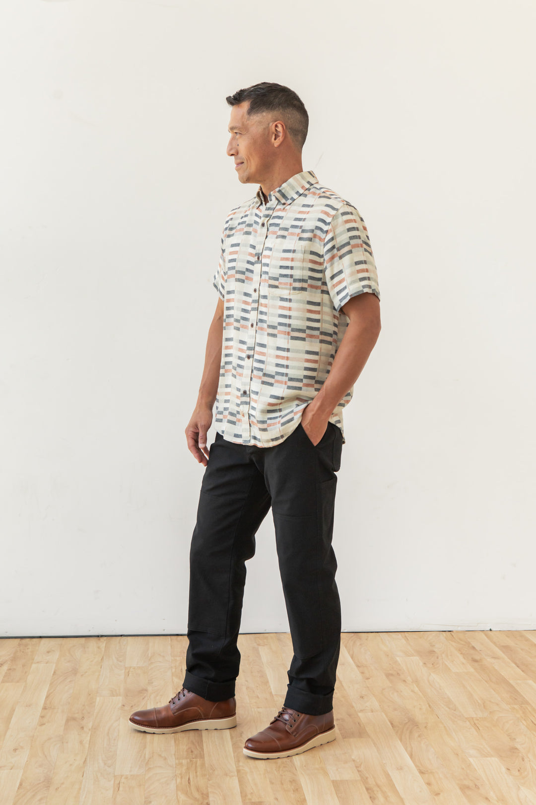 A man stands on a wooden floor against a white wall, wearing the Bridge & Burn Jordan Slim Shirt in Broadway Stripe with black pants and brown shoes—hands in his pockets, looking aside. This shirt is a key piece for contemporary style.