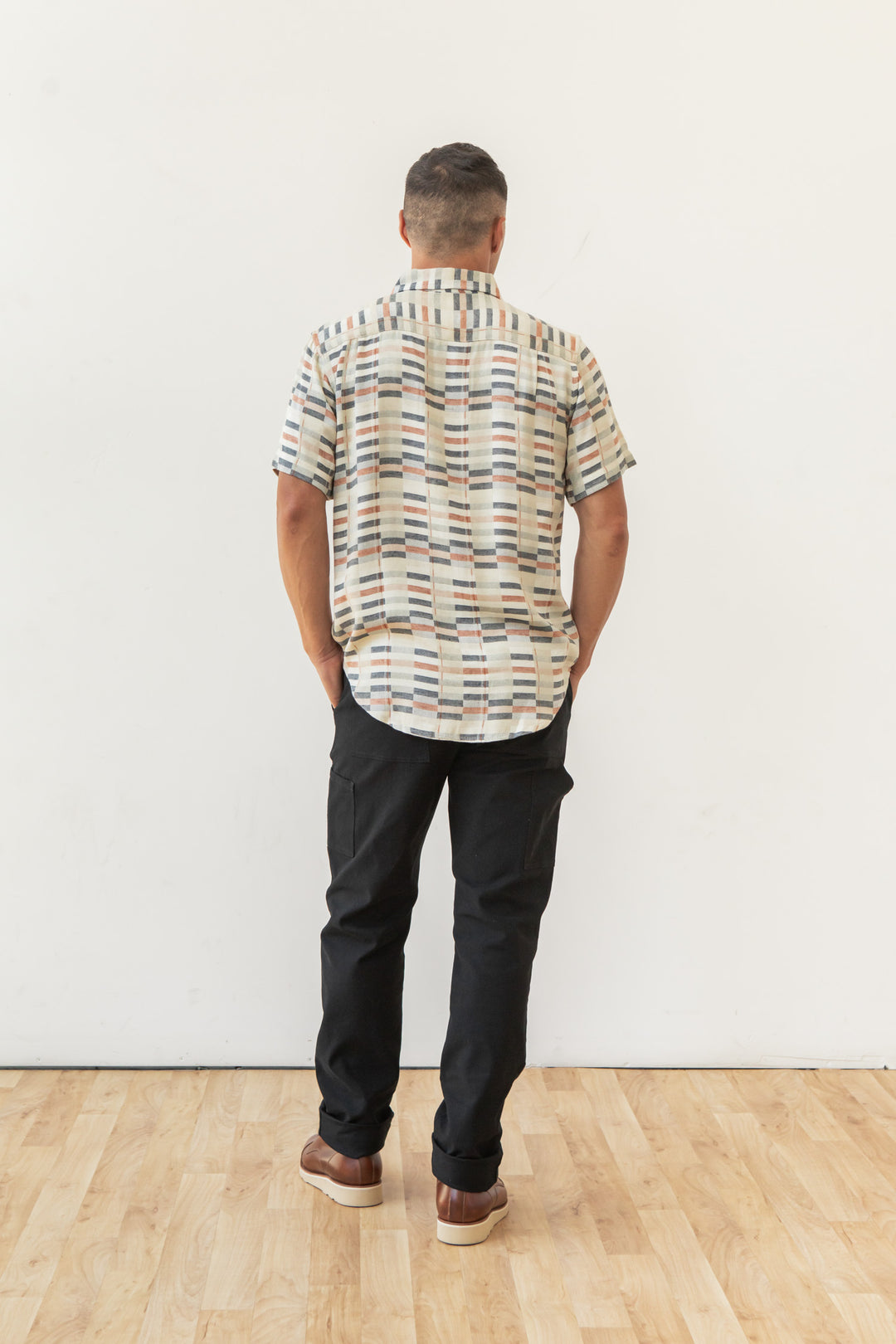 A man stands indoors on a wooden floor facing away from the camera, wearing the Bridge & Burn Jordan Slim Shirt in Broadway Stripe, along with black pants and brown shoes against a plain white wall.