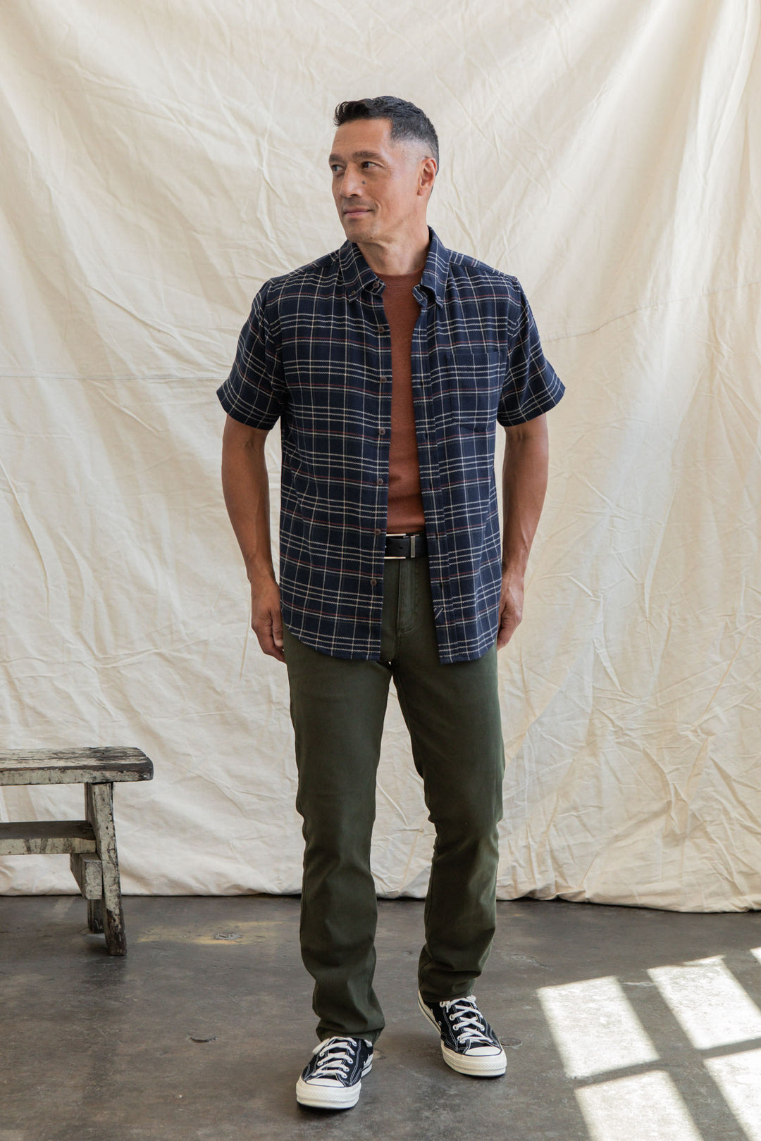 A man stands indoors on a concrete floor, wearing the Bridge & Burn Jordan Slim Shirt in Dusk Herringbone Plaid over a rust T-shirt, olive green pants, and black Converse sneakers. A wooden bench and white draped fabric are in the background.