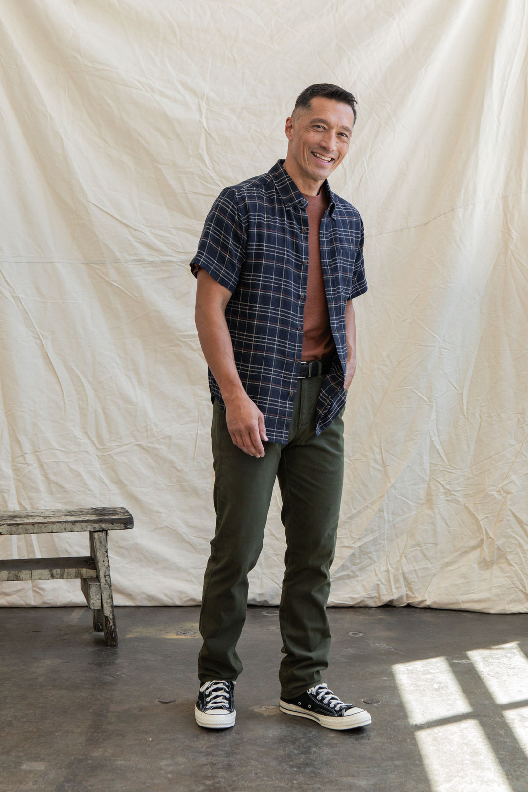 A man wearing the Bridge & Burn Jordan Slim Shirt in Dusk Herringbone Plaid stands smiling in front of a white fabric backdrop beside a wooden bench, sunlight casting patterns on the concrete floor.