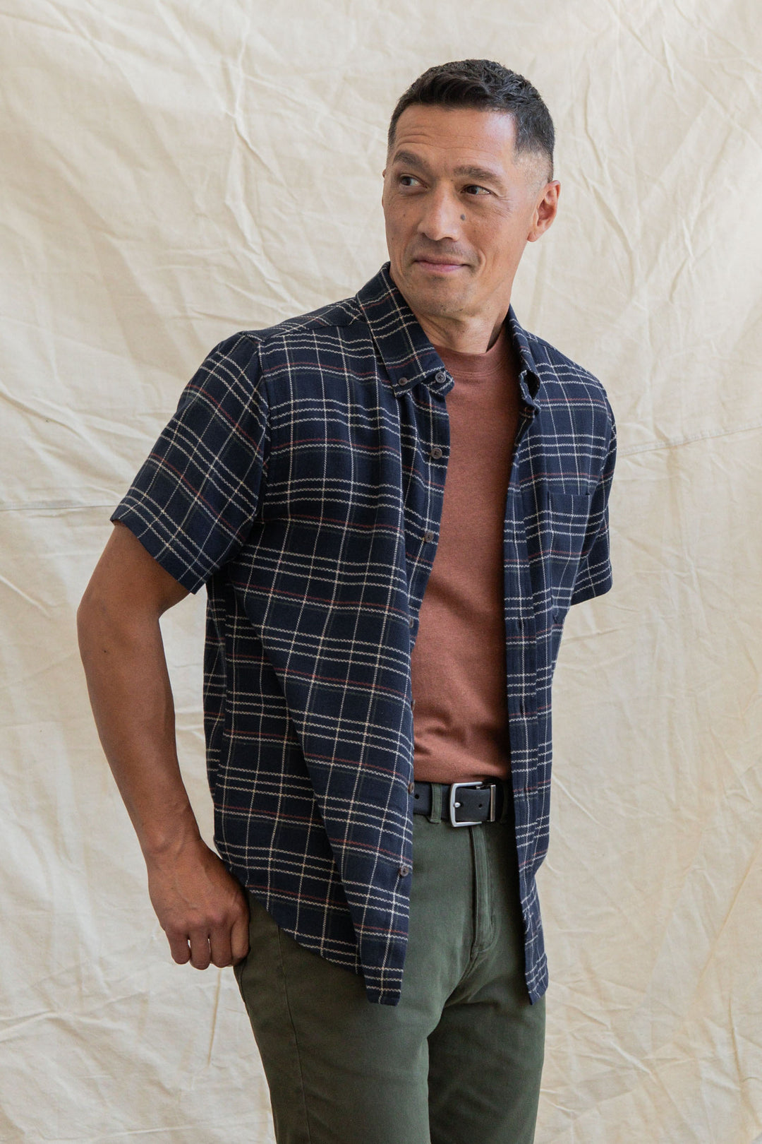 A man stands before a light beige fabric backdrop, wearing the Bridge & Burn Jordan Slim Shirt in Dusk Herringbone Plaid over a brown tee, paired with olive green pants and a black belt. He looks slightly to his right and smiles subtly.