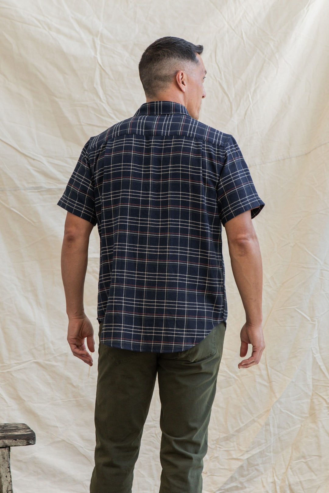 A man faces away from the camera wearing the Bridge & Burn Jordan Slim Shirt in Dusk Herringbone Plaid and olive green pants, standing before a light, wrinkled fabric backdrop.