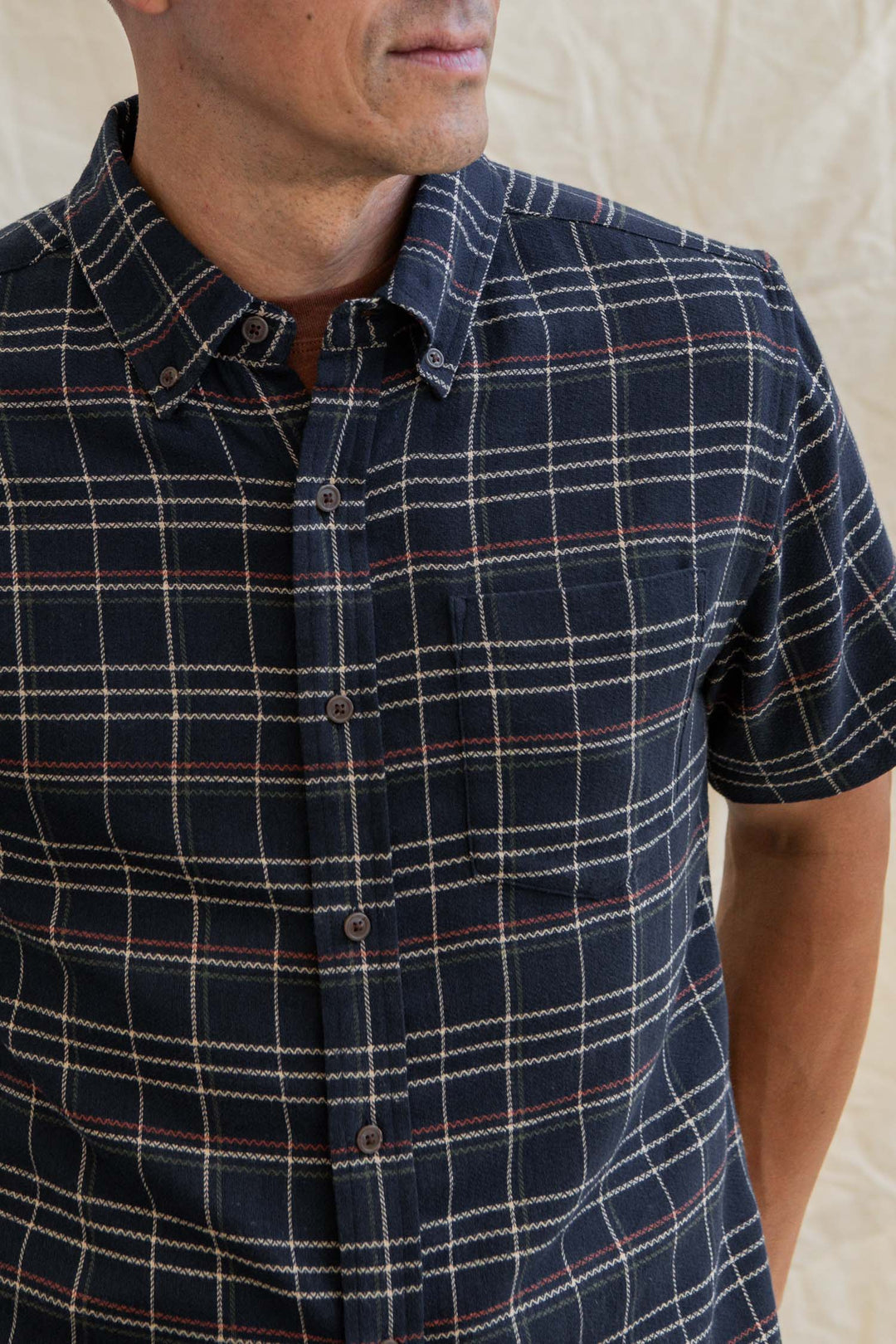 A man stands against a neutral background, wearing the Bridge & Burn Jordan Slim Shirt in Dusk Herringbone Plaid—a modern, short-sleeve button-up with navy, white, and red plaid. His face is partially out of frame.