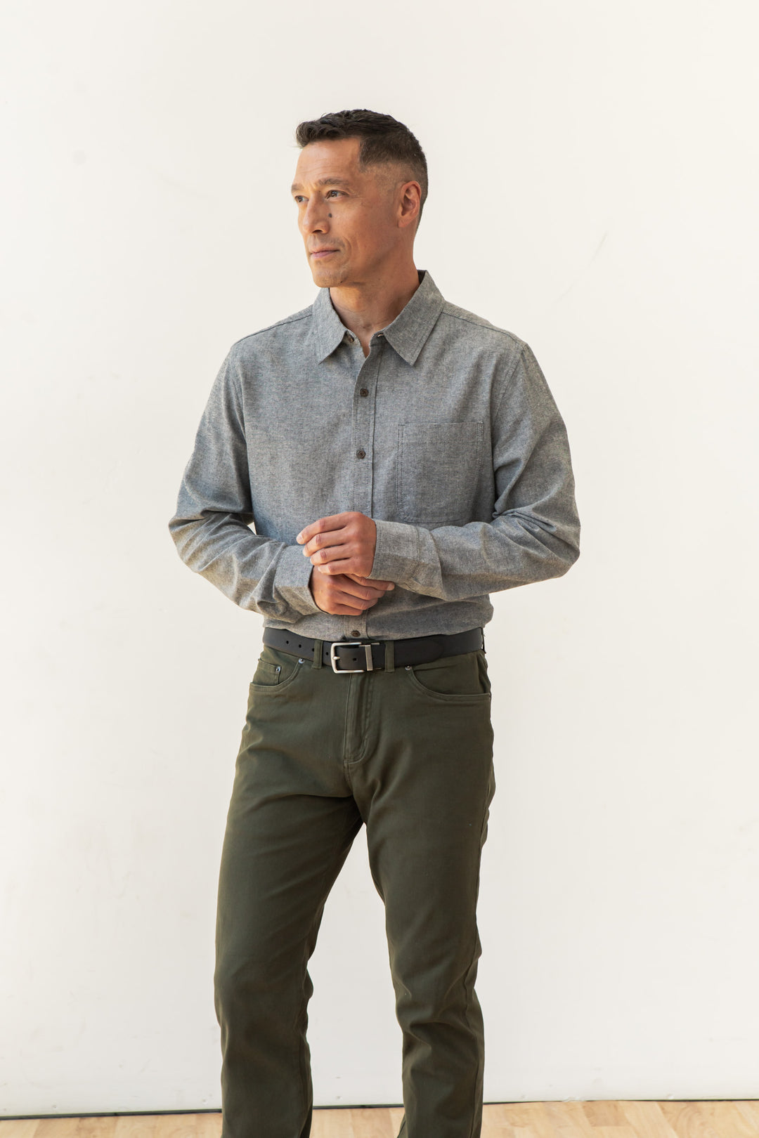 A man stands against a plain white background, wearing the Bridge & Burn Larson Shirt in Iron Chambray and olive green pants. He looks to the side with his hands loosely clasped in front of him.