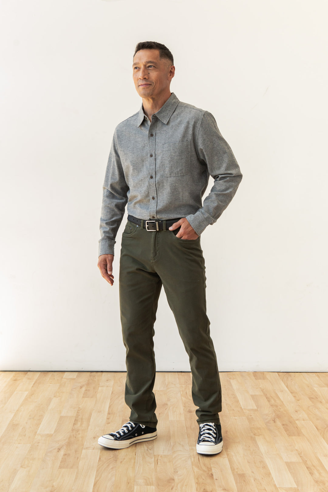 A man with short hair stands indoors on a wooden floor, wearing the Bridge & Burn Larson Shirt in Iron Chambray, dark green pants, a black belt, and black & white sneakers. He looks slightly to the side against a plain white background.