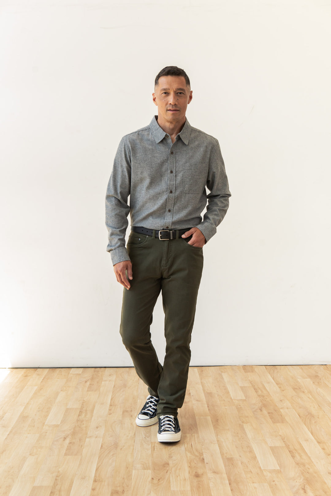 A man with short dark hair and a neutral expression stands on a light wood floor against a white wall, wearing the Bridge & Burn Larson Shirt in Iron Chambray, olive green pants, a black belt, and black-and-white sneakers.