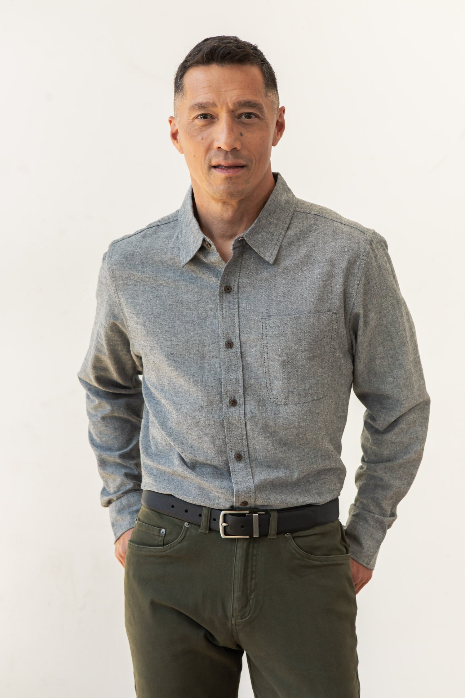 A man with short dark hair stands against a plain light background, wearing the Bridge & Burn Larson Shirt in Iron Chambray with olive green pants and a black belt. One hand is in his pocket as he looks at the camera.