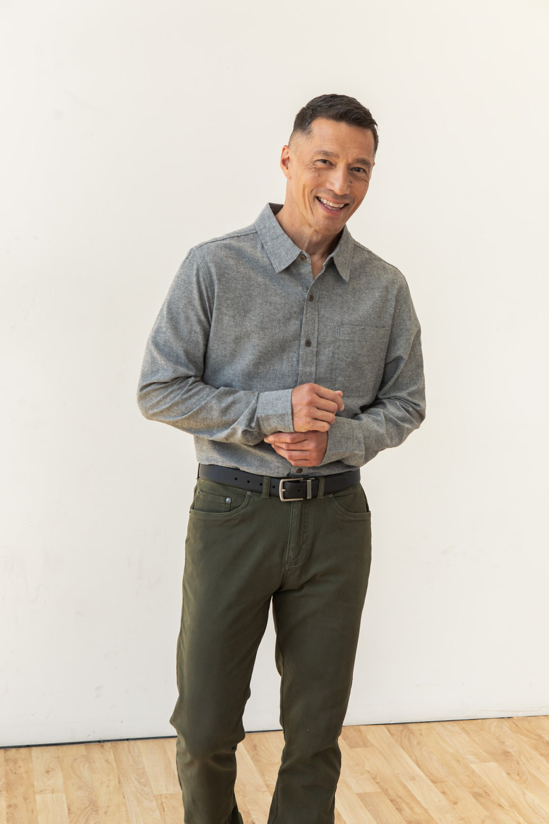 A man with short dark hair stands indoors against a white wall, smiling. He wears the Bridge & Burn Larson Shirt in Iron Chambray tucked into olive green pants with a black belt, hands clasped in front of him.