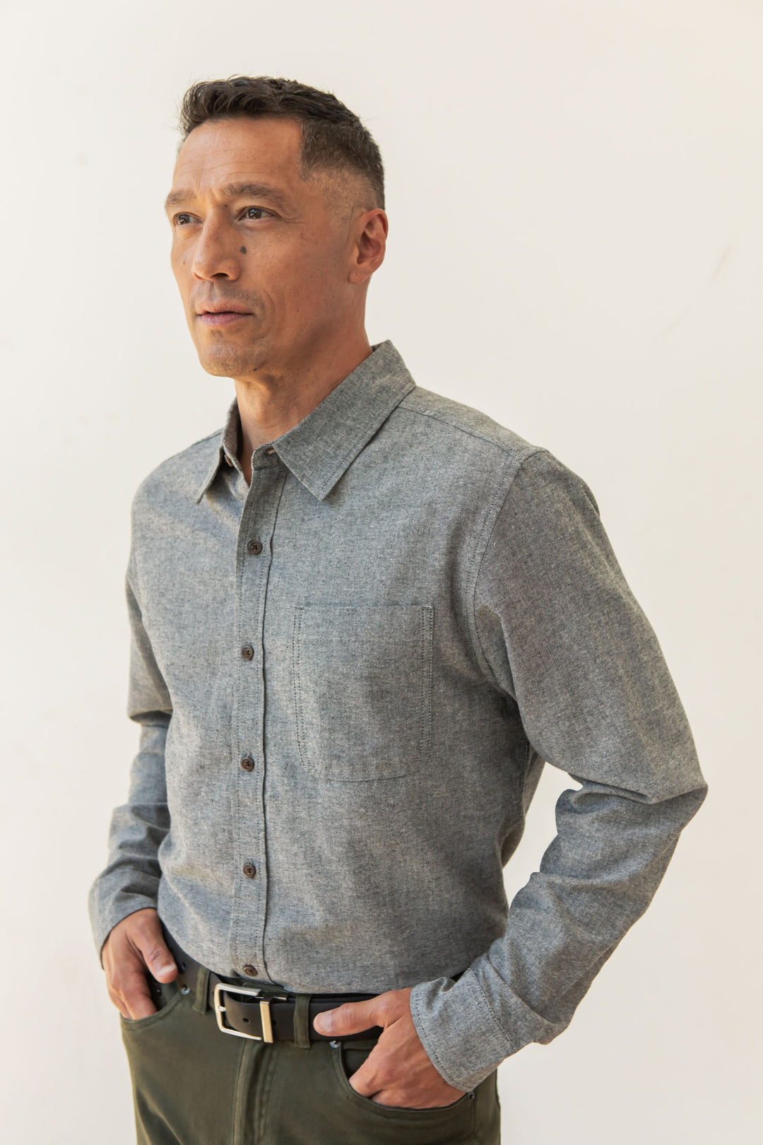 A man with short dark hair stands with his hands in his pockets against a plain light background, wearing the Bridge & Burn Larson Shirt in Iron Chambray and green pants.