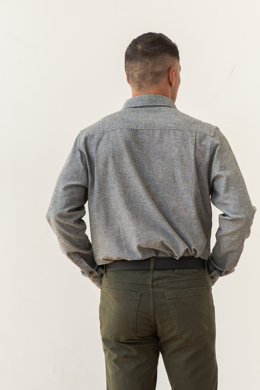 A man stands with his back to the camera, hands on hips, wearing the Bridge & Burn Larson Shirt in Iron Chambray and olive green pants against a plain light background.