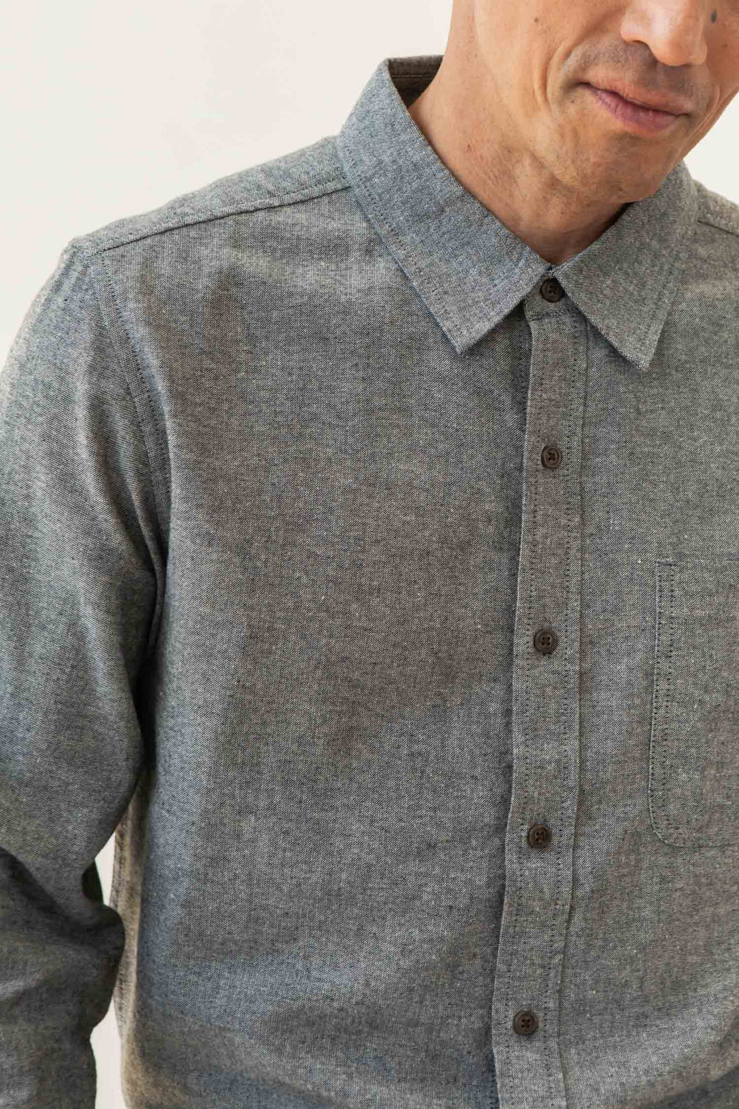 A man wears the Bridge & Burn Larson Shirt in Iron Chambray, a classic fit grey button-up made from recycled cotton. Shown from shoulders to mid-torso, his face is partially visible with a neutral expression.