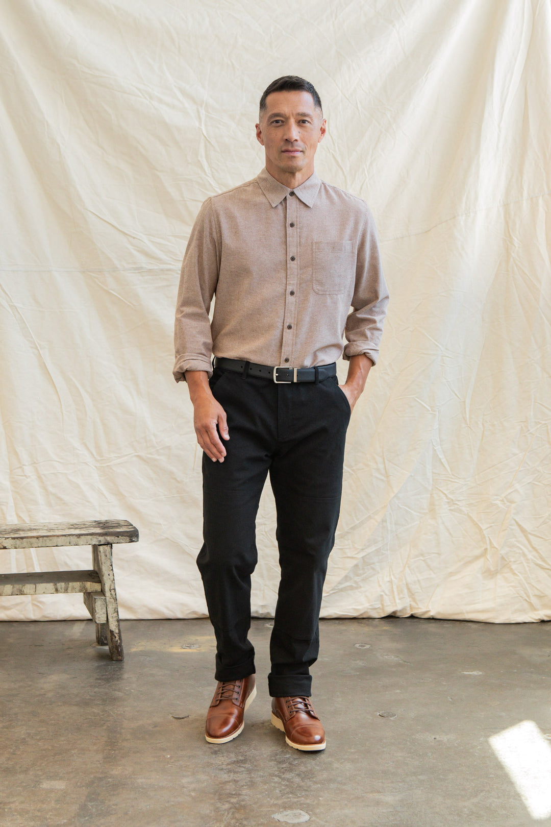 A man stands indoors before a cream fabric backdrop, wearing the Bridge & Burn Larson Shirt in rust chambray, black pants with a black belt, and brown shoes. One hand in his pocket, he stands next to a wooden bench.