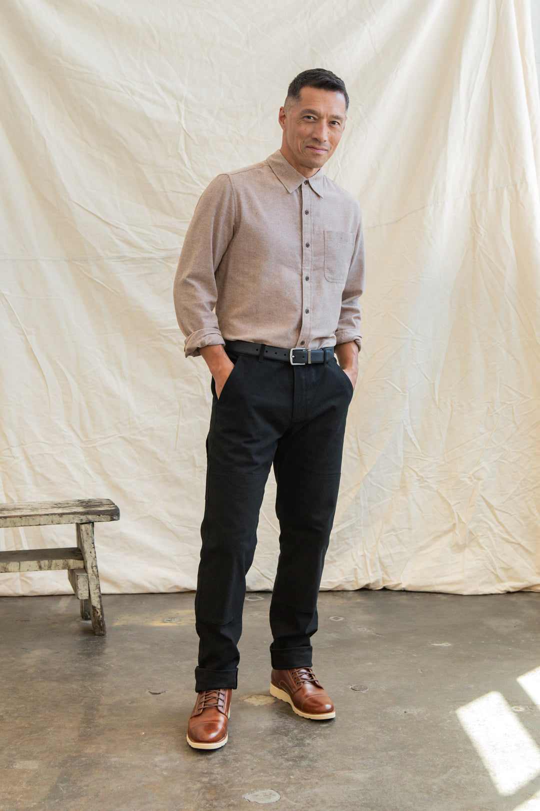 A man stands indoors with his hands in his pockets, wearing the Bridge & Burn Larson Shirt in Rust Chambray, black pants, and brown boots. A white fabric backdrop and wooden stool are visible behind him.