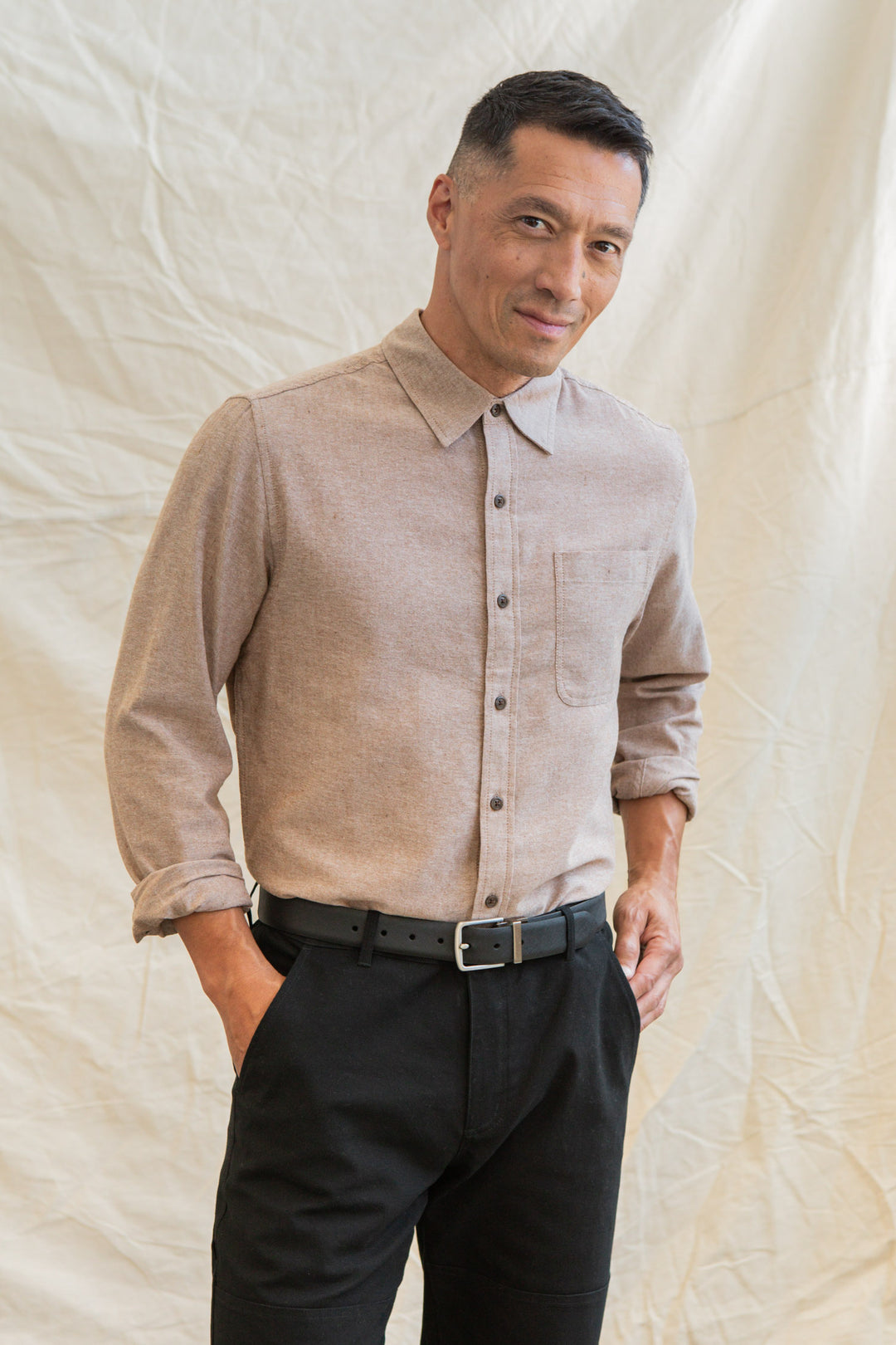 A man with short black hair stands before a light fabric backdrop, smiling gently. He wears the Bridge & Burn Larson Shirt in Rust Chambray, classic fit and recycled cotton, paired with black pants and a belt, one hand in his pocket.