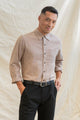 A man with short black hair stands before a light fabric backdrop, smiling gently. He wears the Bridge & Burn Larson Shirt in Rust Chambray, classic fit and recycled cotton, paired with black pants and a belt, one hand in his pocket.