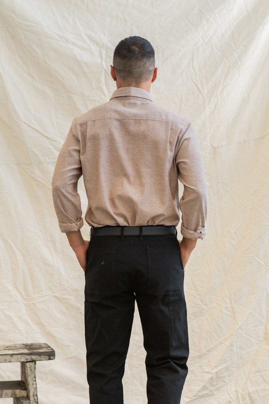 A man with short hair faces away from the camera, wearing the Bridge & Burn Larson Shirt in Rust Chambray and black pants, standing before a cream fabric backdrop. A wooden bench is partially visible on the left.