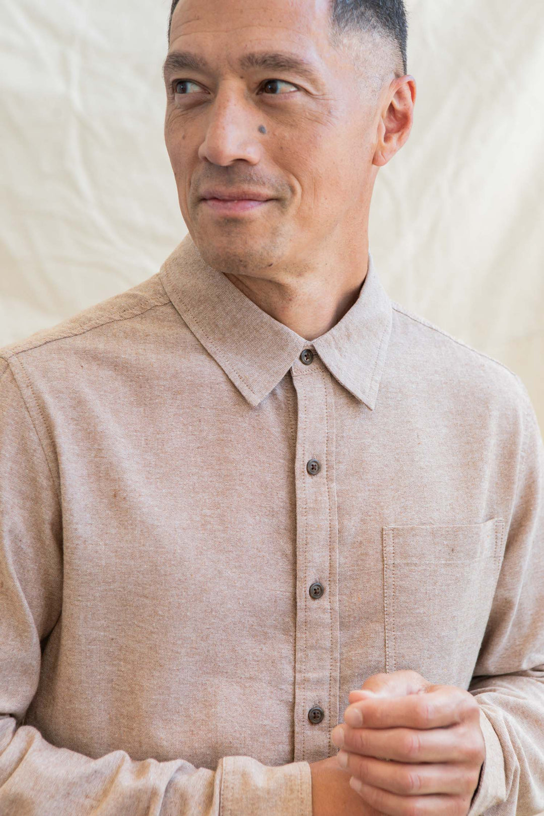 A man in the Bridge & Burn Larson Shirt in Rust Chambray, a classic fit button-up, stands before a light textured background, gazing slightly left with a neutral expression.