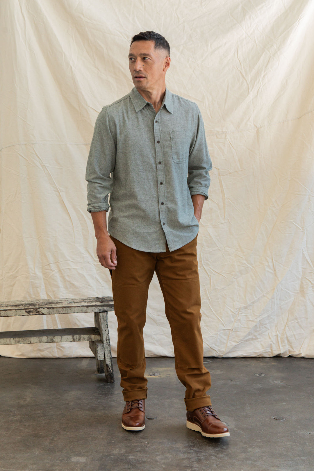 A man stands indoors on a concrete floor, wearing the Bridge & Burn Larson Shirt in Sage Chambray, brown pants, and brown shoes. He looks to the side with one hand in his pocket; a wooden bench and beige backdrop are behind him.