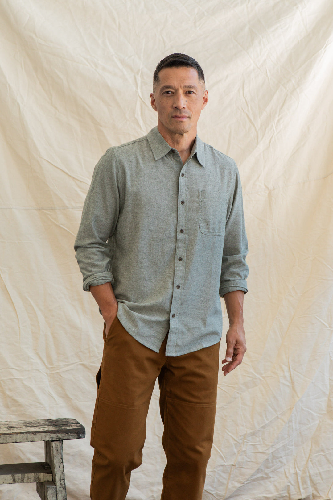 A man stands before a cream textured fabric backdrop, wearing the Bridge & Burn Larson Shirt in Sage Chambray with brown pants and one hand in his pocket. A rustic wooden stool sits partly visible at his side.