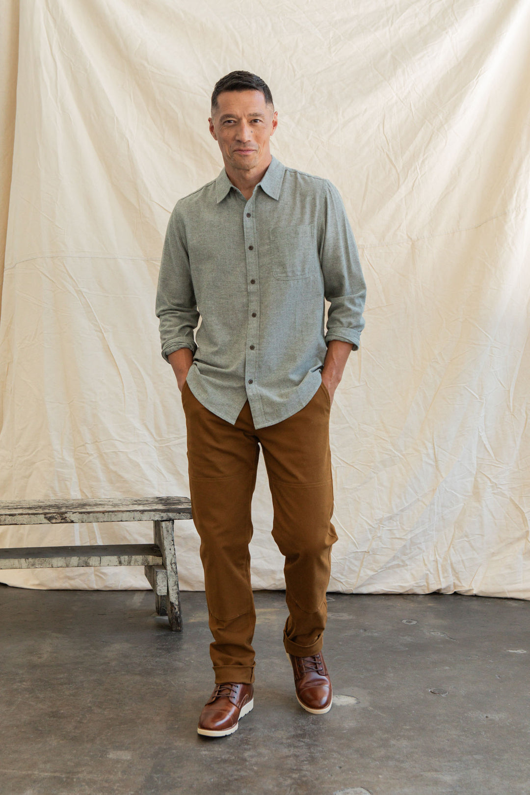 A man stands indoors before a cream backdrop, wearing the Bridge & Burn Larson Shirt in Sage Chambray, brown pants, and brown boots. His hands are in his pockets and he has a relaxed expression.