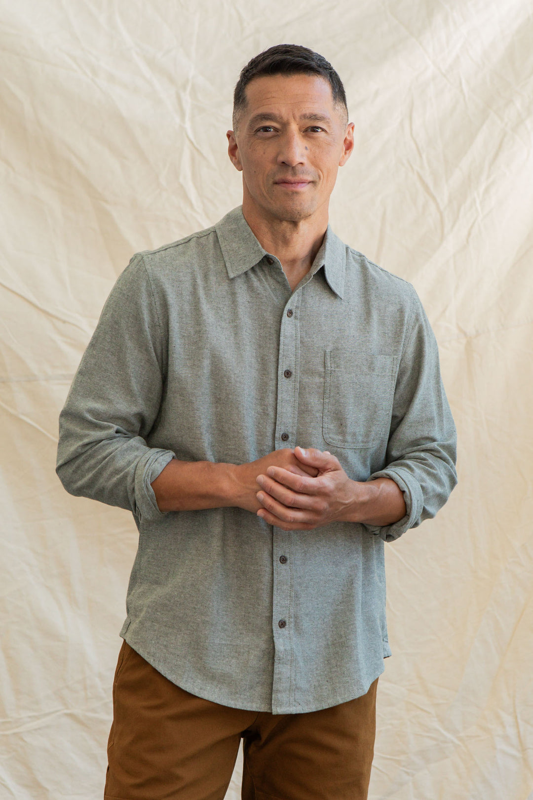 A man with short dark hair, wearing the Bridge & Burn Larson Shirt in Sage Chambray and brown pants, stands before a crumpled fabric backdrop, looking at the camera with a gentle expression and his hands loosely clasped.