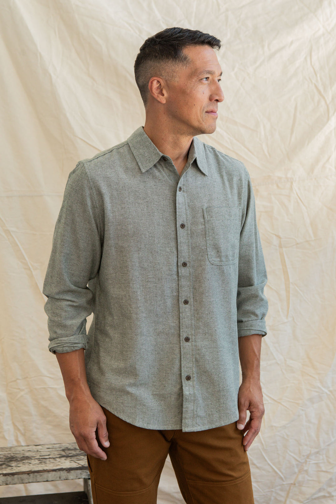 A man with short hair, wearing a Bridge & Burn Larson Shirt in Sage Chambray and brown pants, stands before a beige fabric backdrop, looking to his left with a neutral expression.