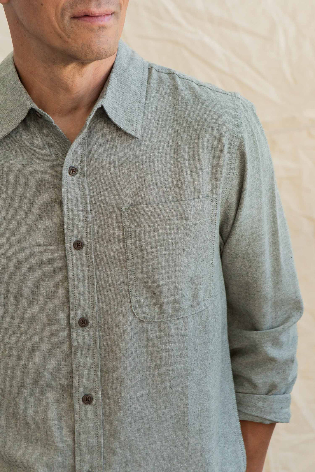 A person wears the Bridge & Burn Larson Shirt in Sage Chambray, sleeves rolled up, standing before a beige background. Only the lower half of their face and upper torso are shown.