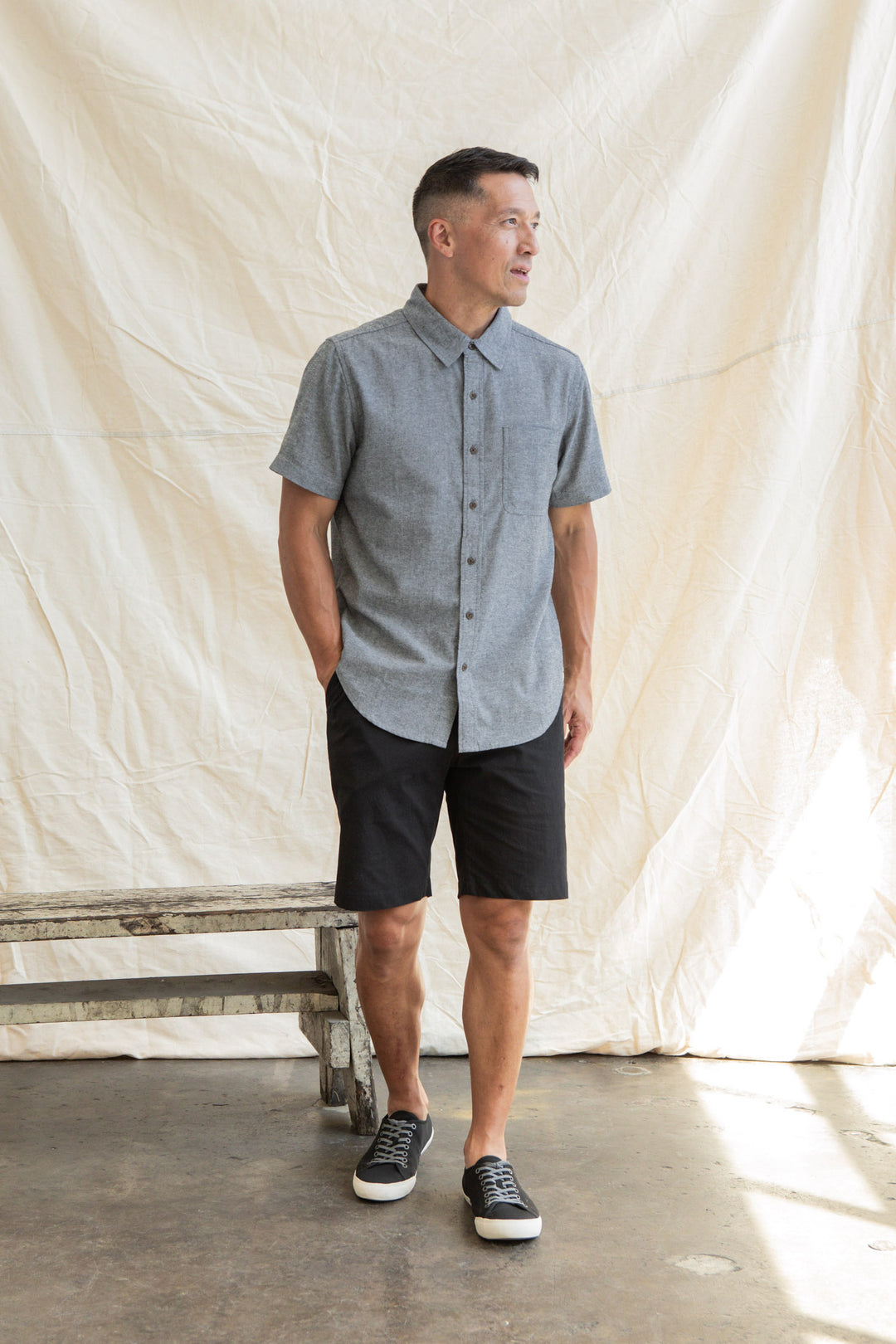 A man stands indoors before a cream draped backdrop, wearing the Bridge & Burn Malcolm Shirt in Iron Chambray, black shorts, and black sneakers with white soles. Hands in pockets, he looks to the side.