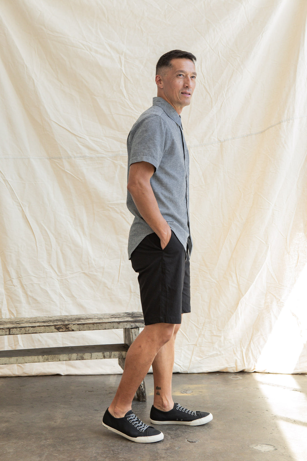 A man stands sideways with his hands in the pockets of black shorts, wearing the Bridge & Burn Malcolm Shirt in Iron Chambray and black sneakers. He is indoors, posed before a white fabric backdrop.