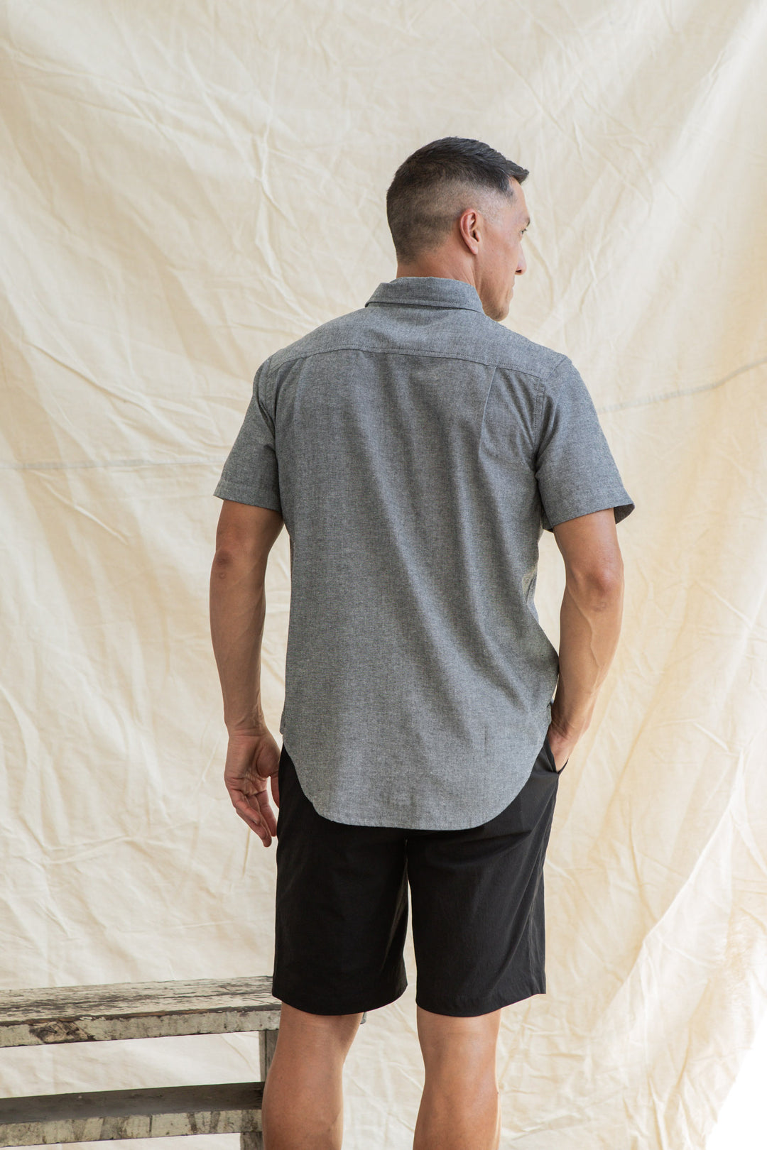 A man stands with his back to the camera, wearing the Bridge & Burn Malcolm Shirt in Iron Chambray and black shorts, one hand in his pocket. A neutral fabric backdrop hangs behind him.
