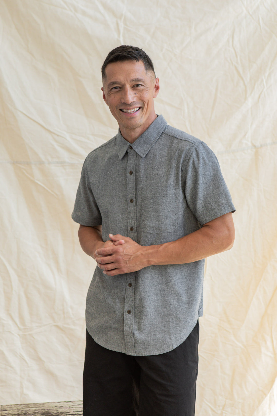 A man with short dark hair smiles in front of a light fabric backdrop, wearing the Bridge & Burn Malcolm Shirt in Iron Chambray and black pants, his hands loosely clasped.