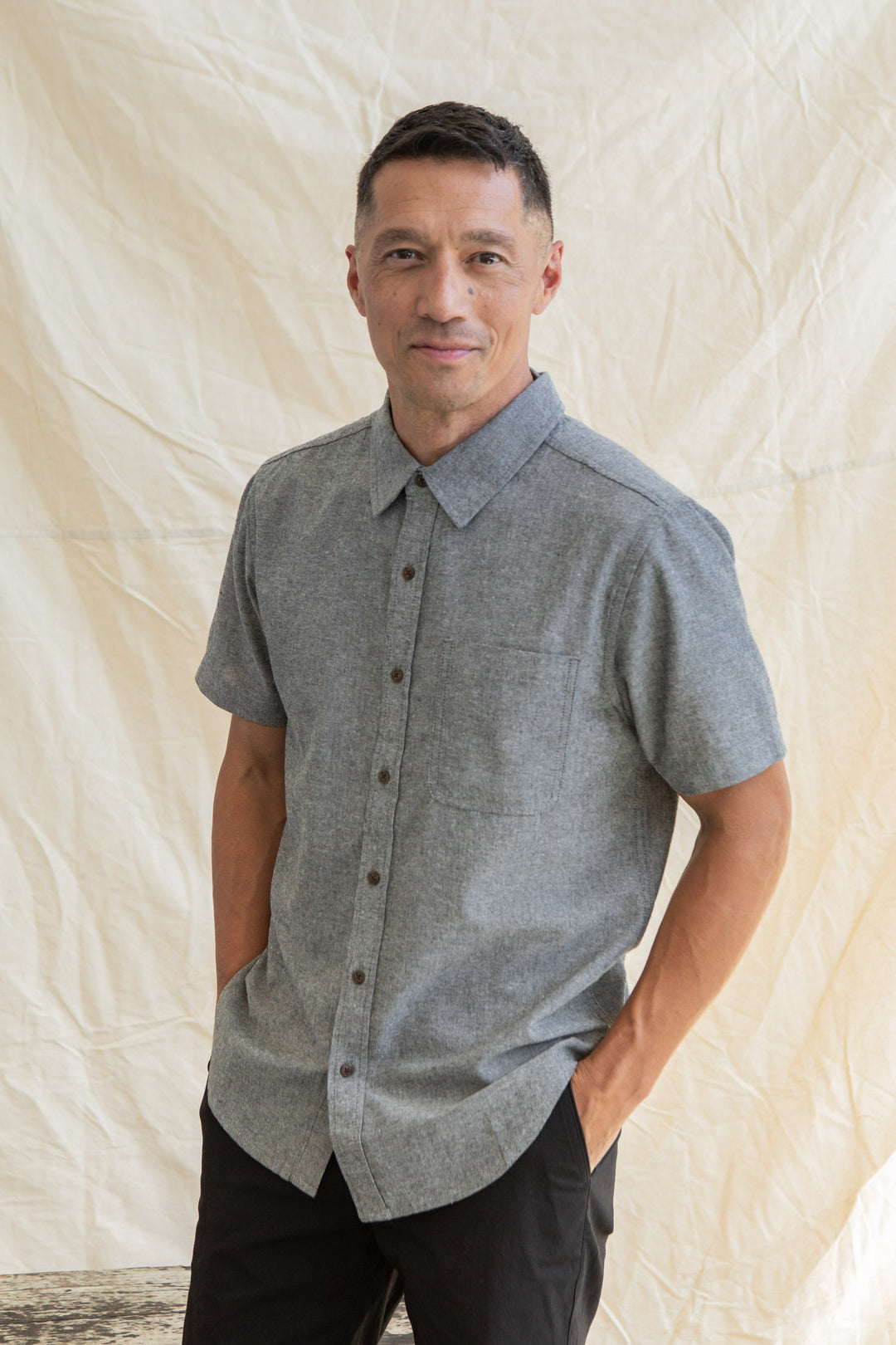 A man stands before a light backdrop, hands in pockets, smiling at the camera while wearing the Bridge & Burn Malcolm Shirt in Iron Chambray and black pants.