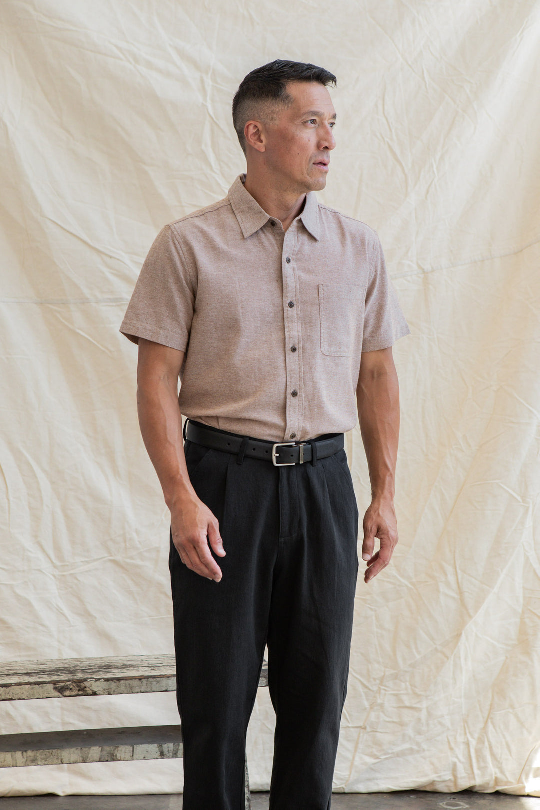 A man stands in front of a beige fabric backdrop wearing the Bridge & Burn Malcolm Shirt in Rust Chambray and black pants, facing slightly right with one arm relaxed at his side.