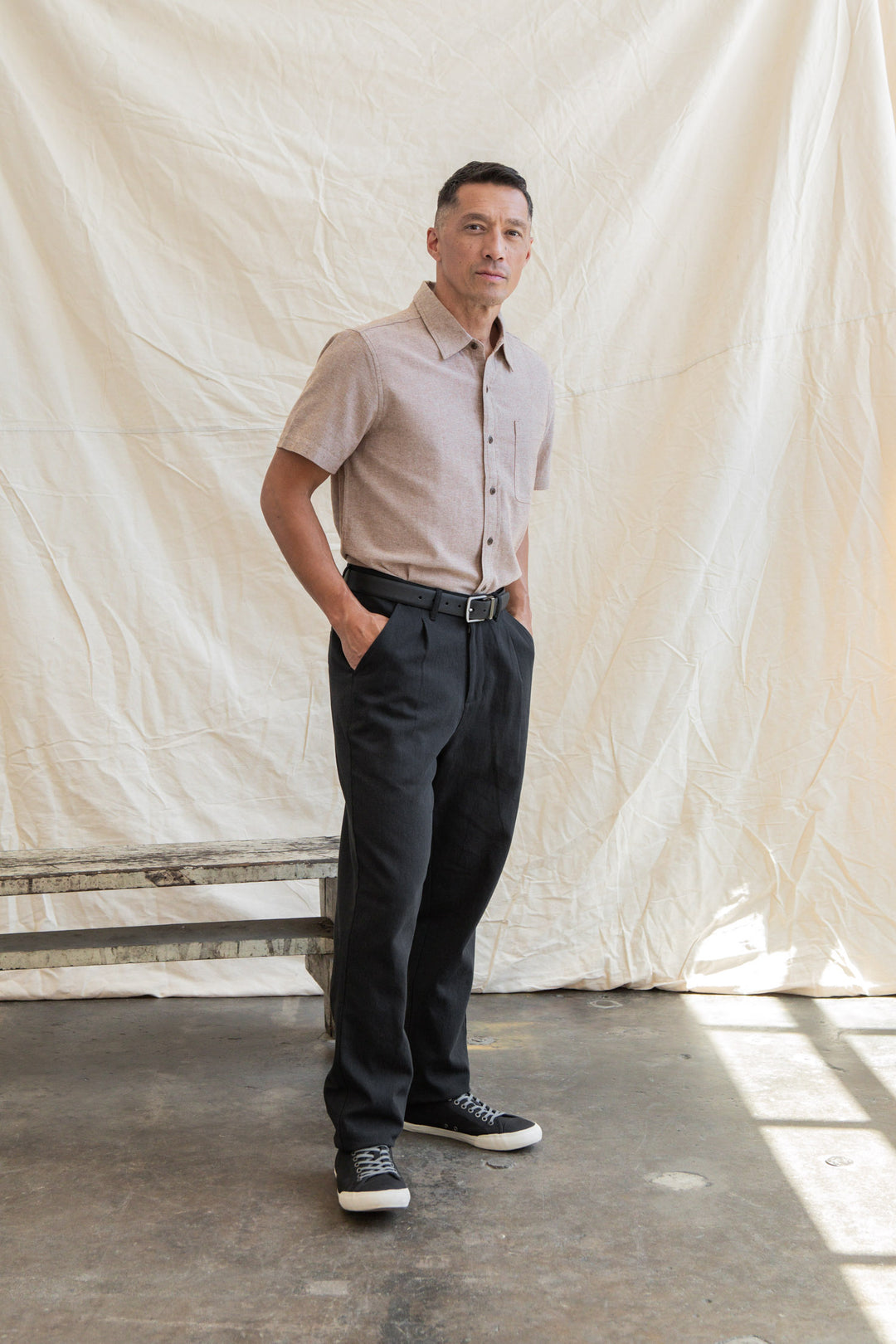 A man stands indoors on concrete, hands in pockets, wearing the Bridge & Burn Malcolm Shirt in Rust Chambray, dark trousers, and black sneakers before a cream-colored fabric backdrop.