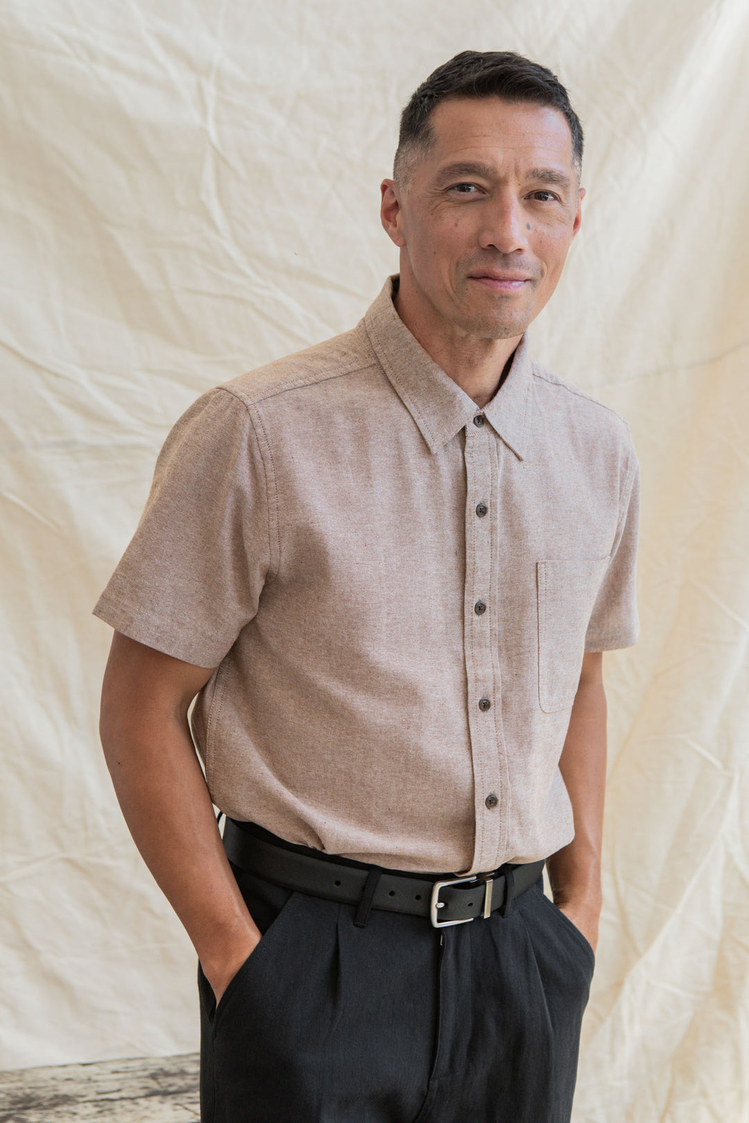 A man with short dark hair wears the Bridge & Burn Malcolm Shirt in Rust Chambray and black trousers, standing in front of a neutral fabric backdrop with his hands in his pockets, smiling slightly.