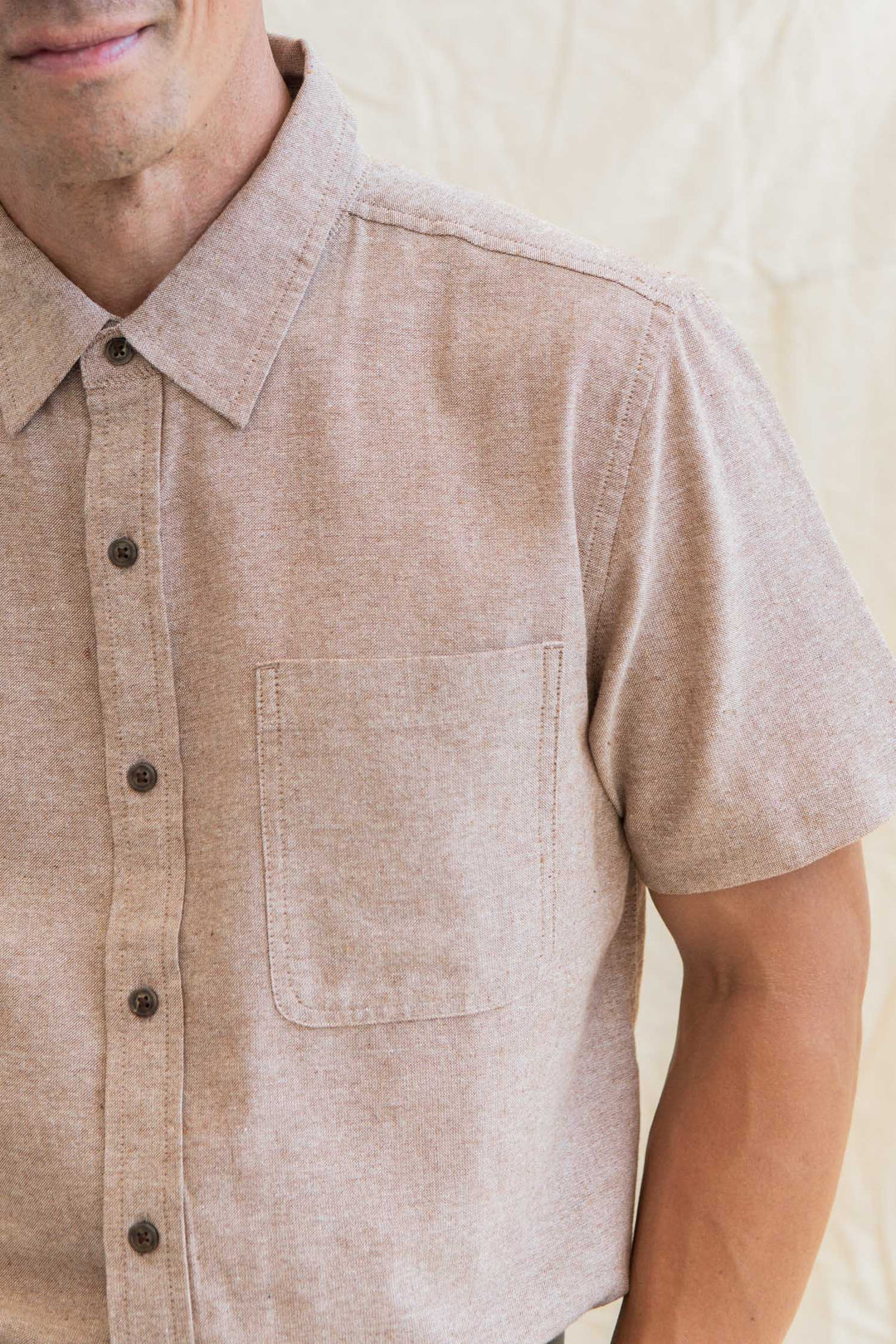 A person wears the Bridge & Burn Malcolm Shirt in Rust Chambray, a light brown short-sleeve button-up with chest pocket, made from recycled cotton, shown from shoulders to waist against a neutral background.