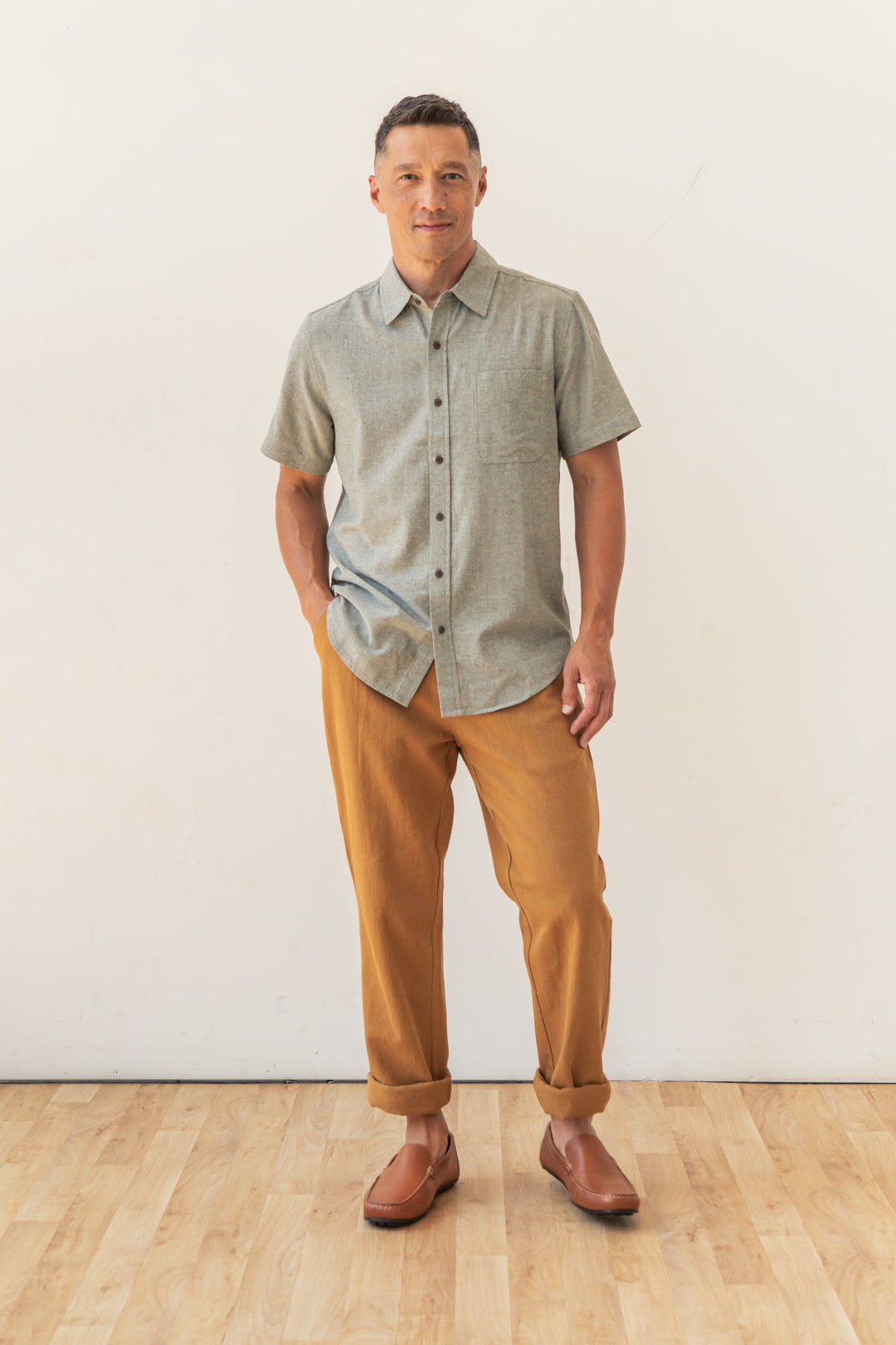A man stands indoors on a light wood floor, wearing the Bridge & Burn Malcolm Shirt in Sage Chambray—a sustainable short sleeve men's button-up—paired with tan cuffed pants and brown slip-on shoes. He looks at the camera, hands in his pockets.
