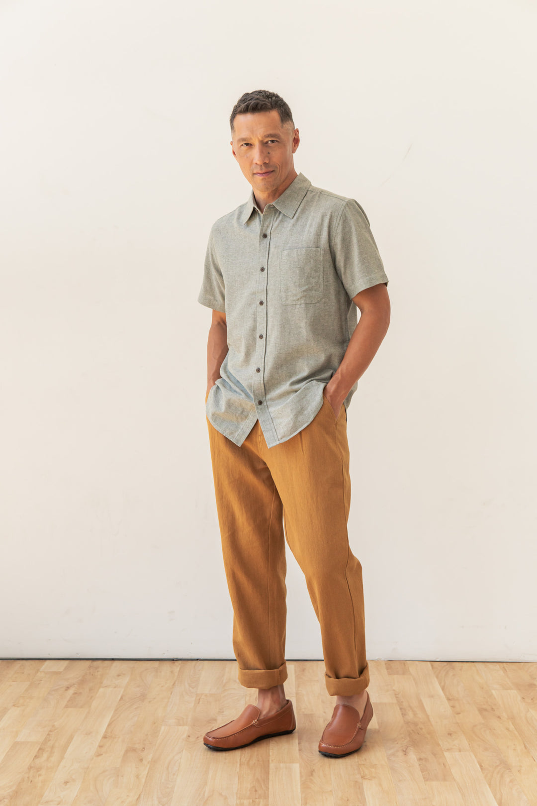 A man stands indoors against a plain white wall, wearing the Bridge & Burn Malcolm Shirt in Sage Chambray, tan cuffed pants, and brown loafers, with his hands in his pockets.
